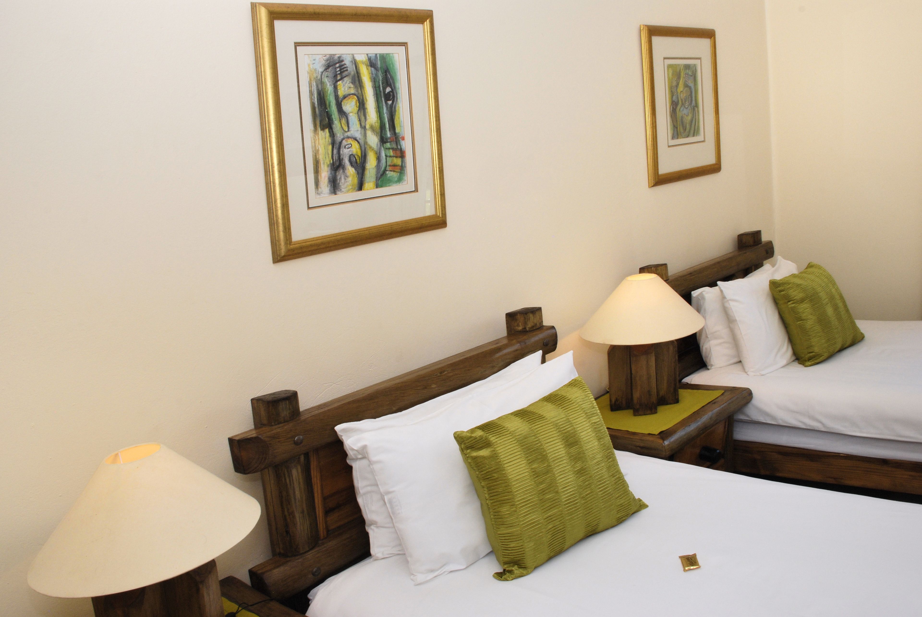 Een hotelkamer met twee bedden, elk met een houten hoofdbord, witte beddengoed en een groen kussen. Boven de bedden hangen twee kunstwerken in een lijst.