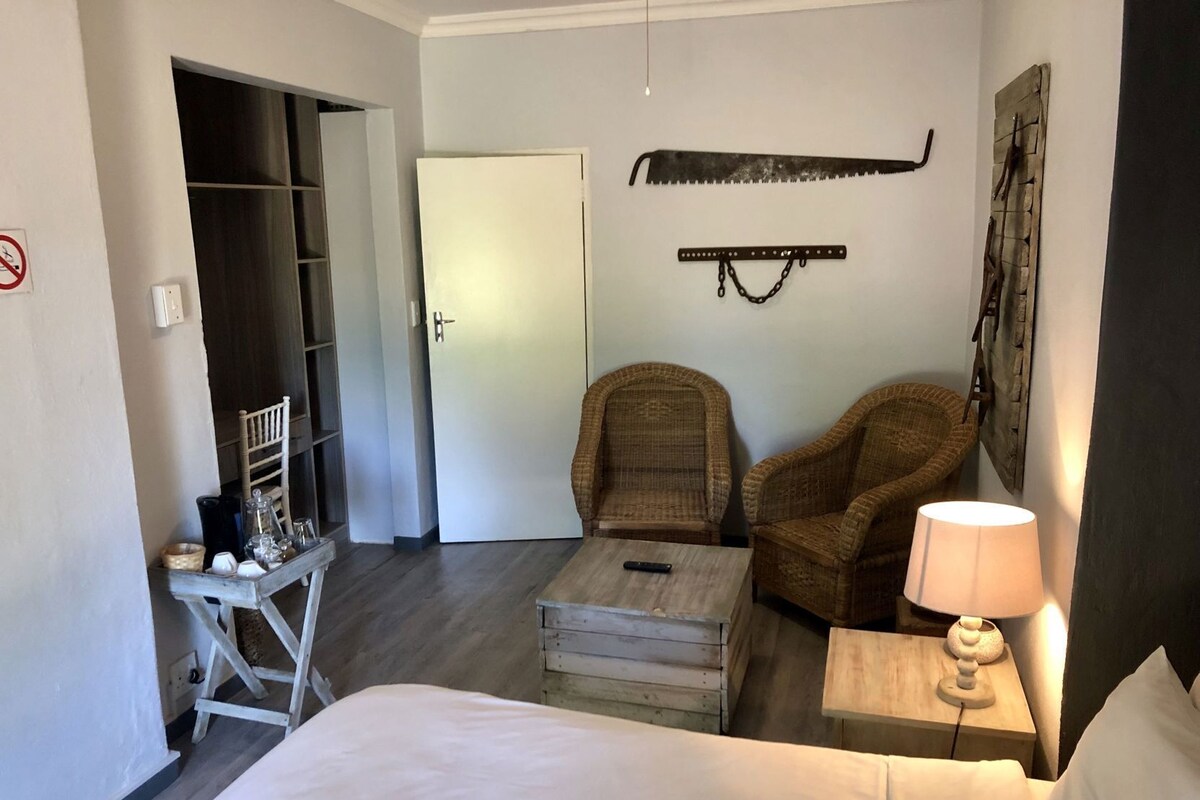 Een slaapkamer met een bed, twee rieten stoelen, een bijzettafeltje, een nachtkastje met een lamp, een houten bankje, een kledingkast en een deur.
