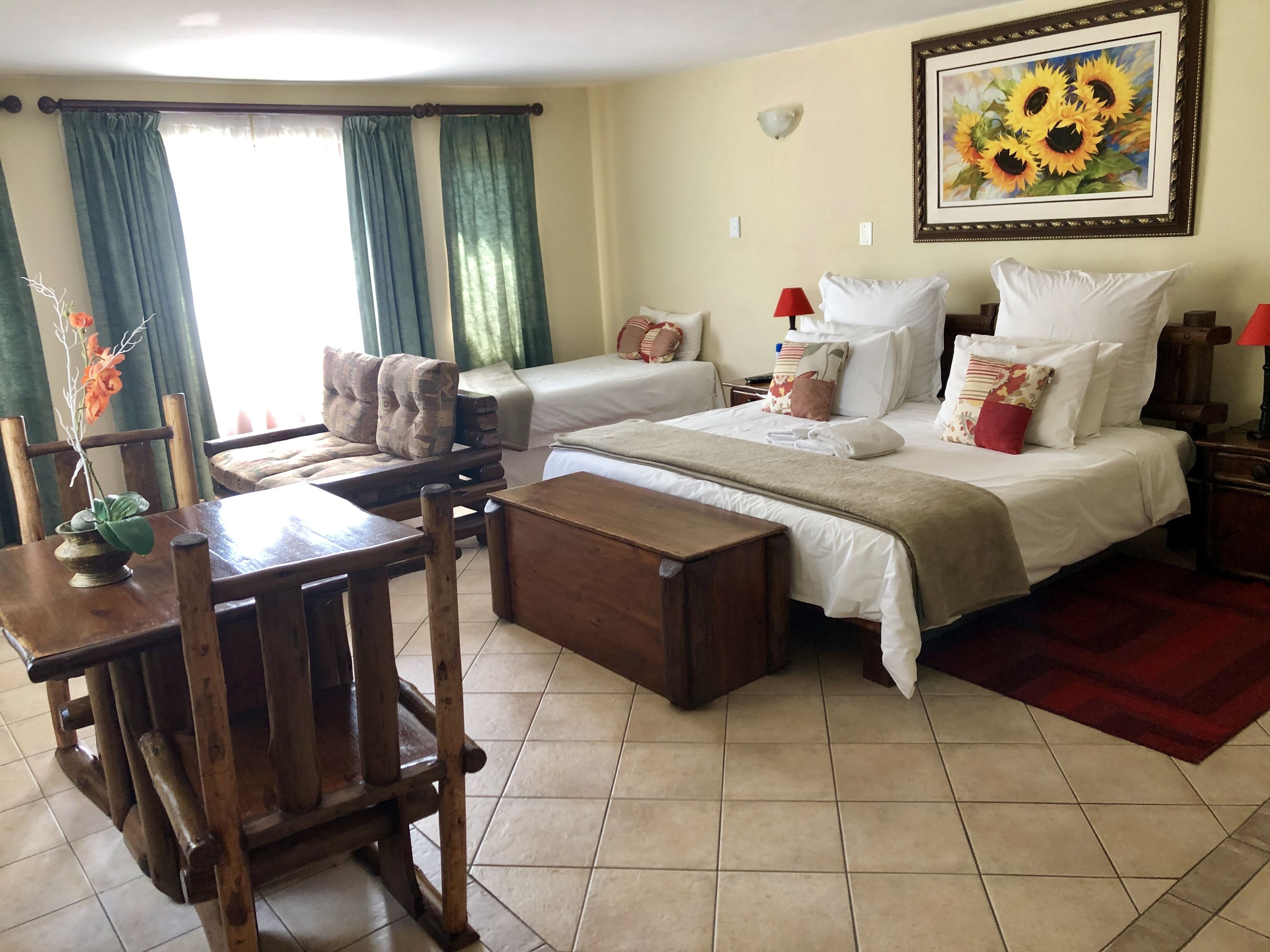 A hotel room with two beds, a dining table, chairs, a small table with a vase, and a painting of sunflowers on the wall.