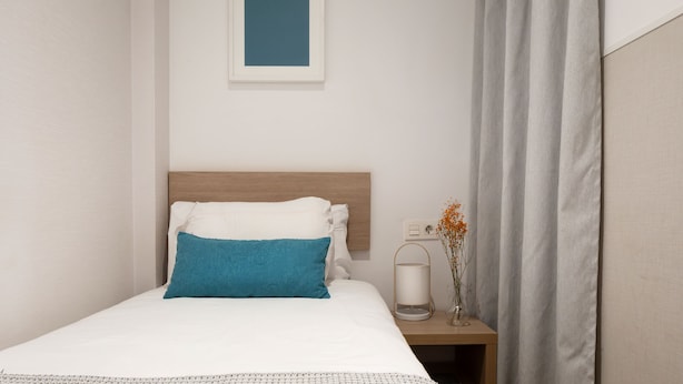 A neatly made bed with a white and blue pillow, a bedside table with a vase of flowers, and a framed picture on the wall. - Feelathome Plaza Apartments (Barcelona)