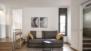A modern living room with a grey sofa, two cushions, a small table with a laptop, and a geometric rug. - Feelathome Plaza Apartments (Barcelona)