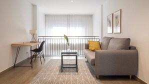 A modern living room with a sofa, a desk, and a chair. There is a window with curtains and a railing. - Feelathome Plaza Apartments (Barcelona)