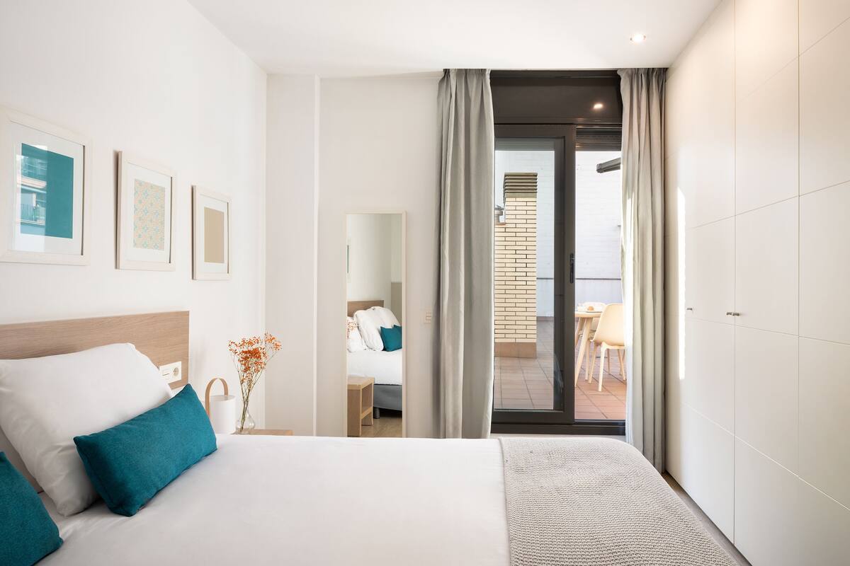 Een moderne hotelkamer met een groot bed, een kleintje tafeltje met een vaas, een balkon met uitzicht en een schuifdeur naar een eetgedeelte.