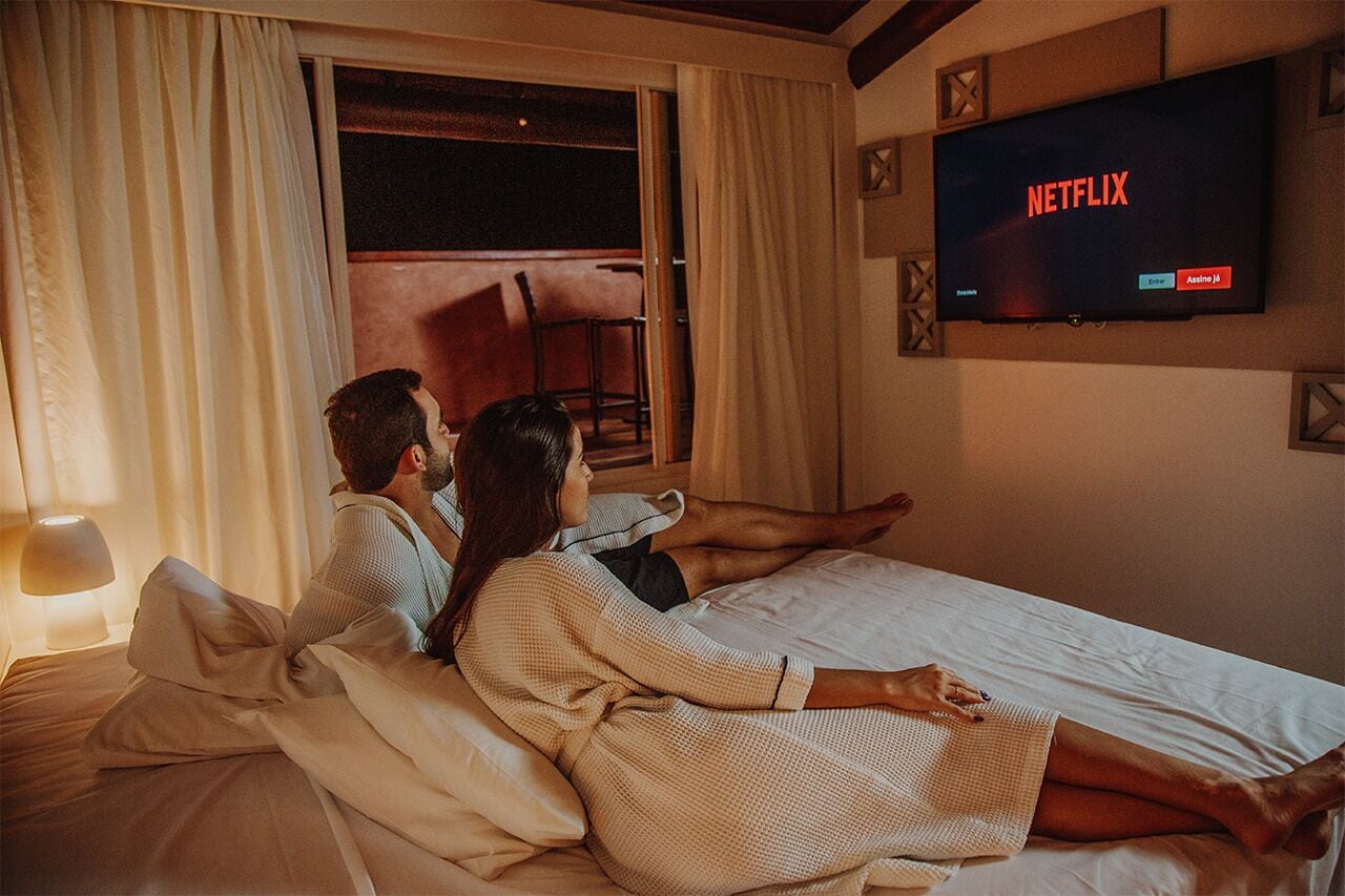 A couple lying on a bed watching TV in a hotel room with a large window and a wall-mounted TV displaying 'NETFLIX'.