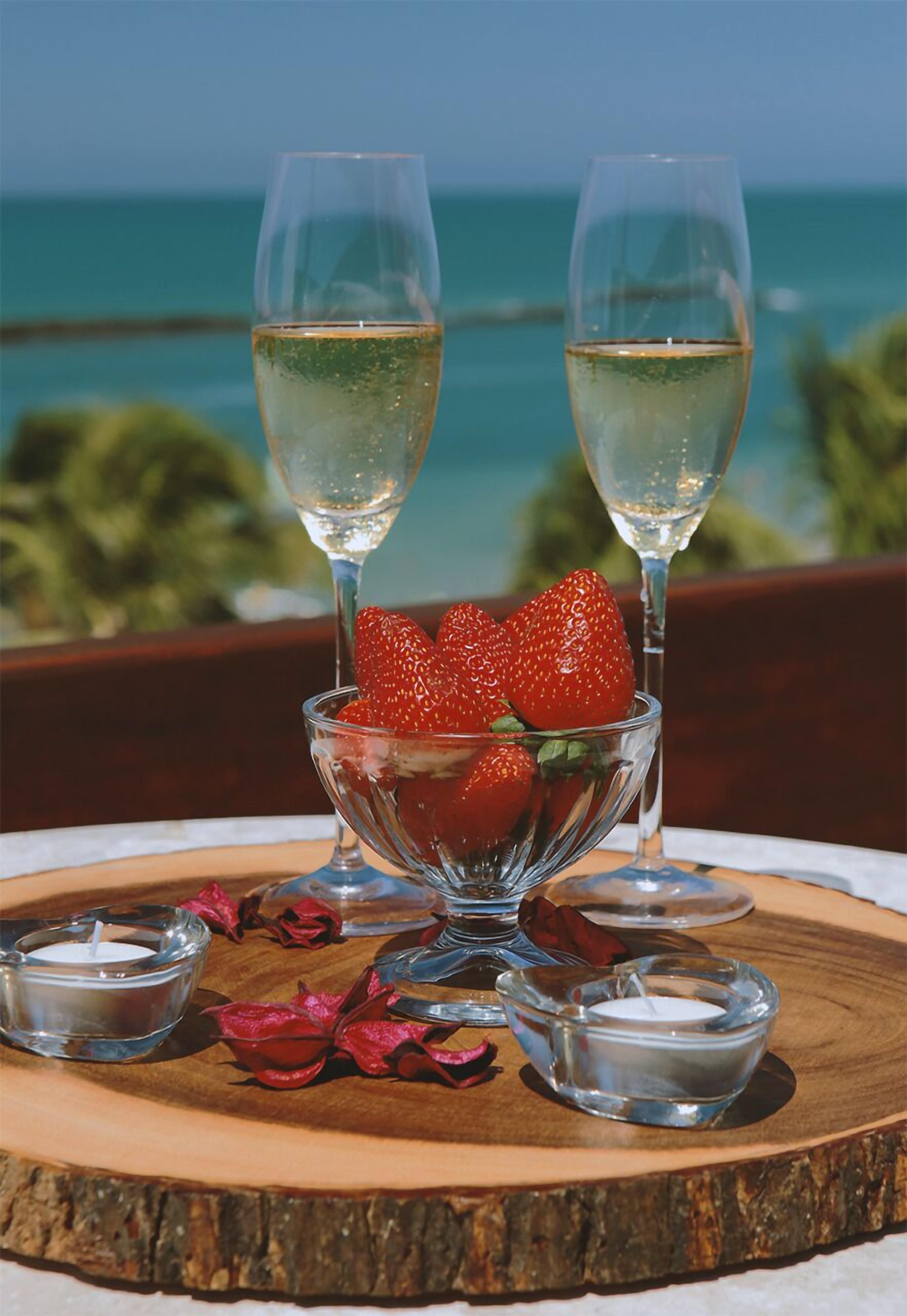 A table with two glasses of white wine, a bowl of strawberries, and small candles on a wooden board with a view of the ocean.