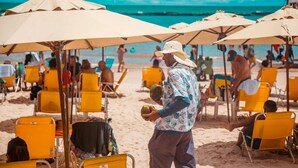 Una playa con sillas y sombrillas amarillas, un hombre sosteniendo una nuez de coco y gente al fondo.