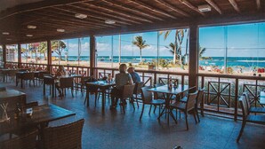 Un área de comedor al aire libre con mesas y sillas, con vista a una playa con palmeras y el océano.