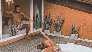 Un hombre y una mujer en una terraza de madera con un jacuzzi, rodeados de plantas en macetas y un edificio con tejado de baldosas.