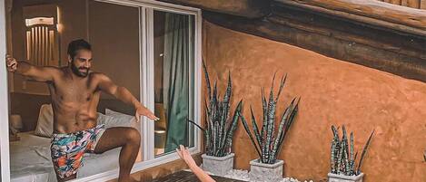 A man and a woman in swimwear on a wooden deck with a hot tub, potted plants, and a building with a tiled roof.