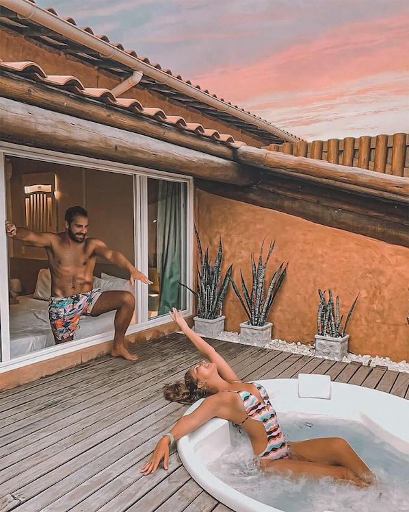 Un hombre y una mujer en una terraza de madera con un jacuzzi, rodeados de plantas en macetas y un edificio con tejado de baldosas.