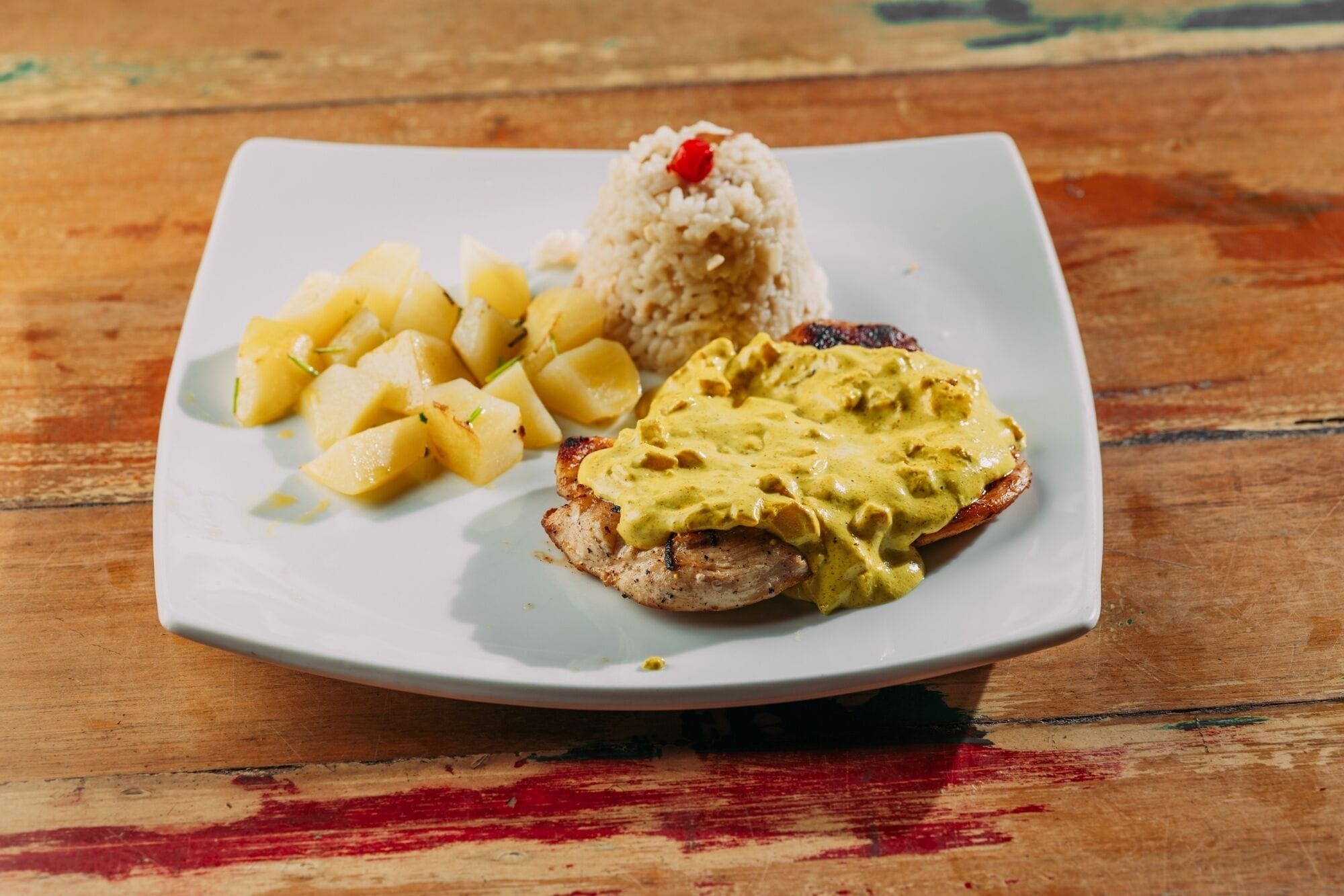 Un plato con carne a la parrilla cubierta con salsa amarilla, acompañamiento de arroz y patatas en cubos.