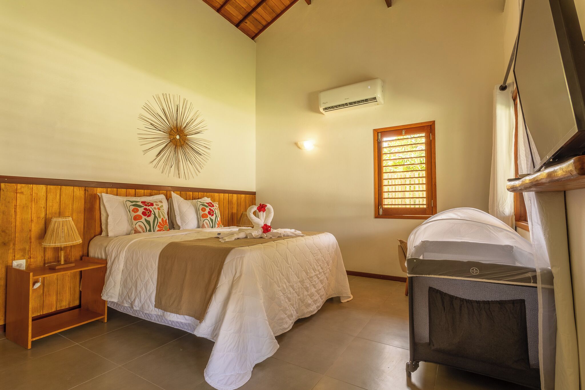 Une chambre bien rangée, avec un lit, une table de chevet, une lampe, des coussins décoratifs, un climatiseur fixé au mur, une fenêtre avec des stores et un ventilateur de plafond.