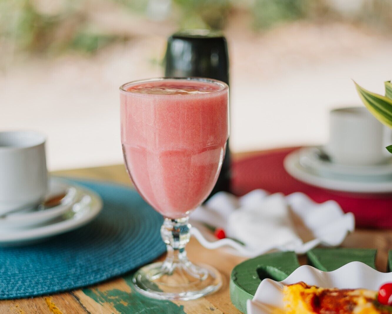 A glass of pink smoothie on a wooden table with a white cup and saucer, a green placemat, and a red plate with a white flower.