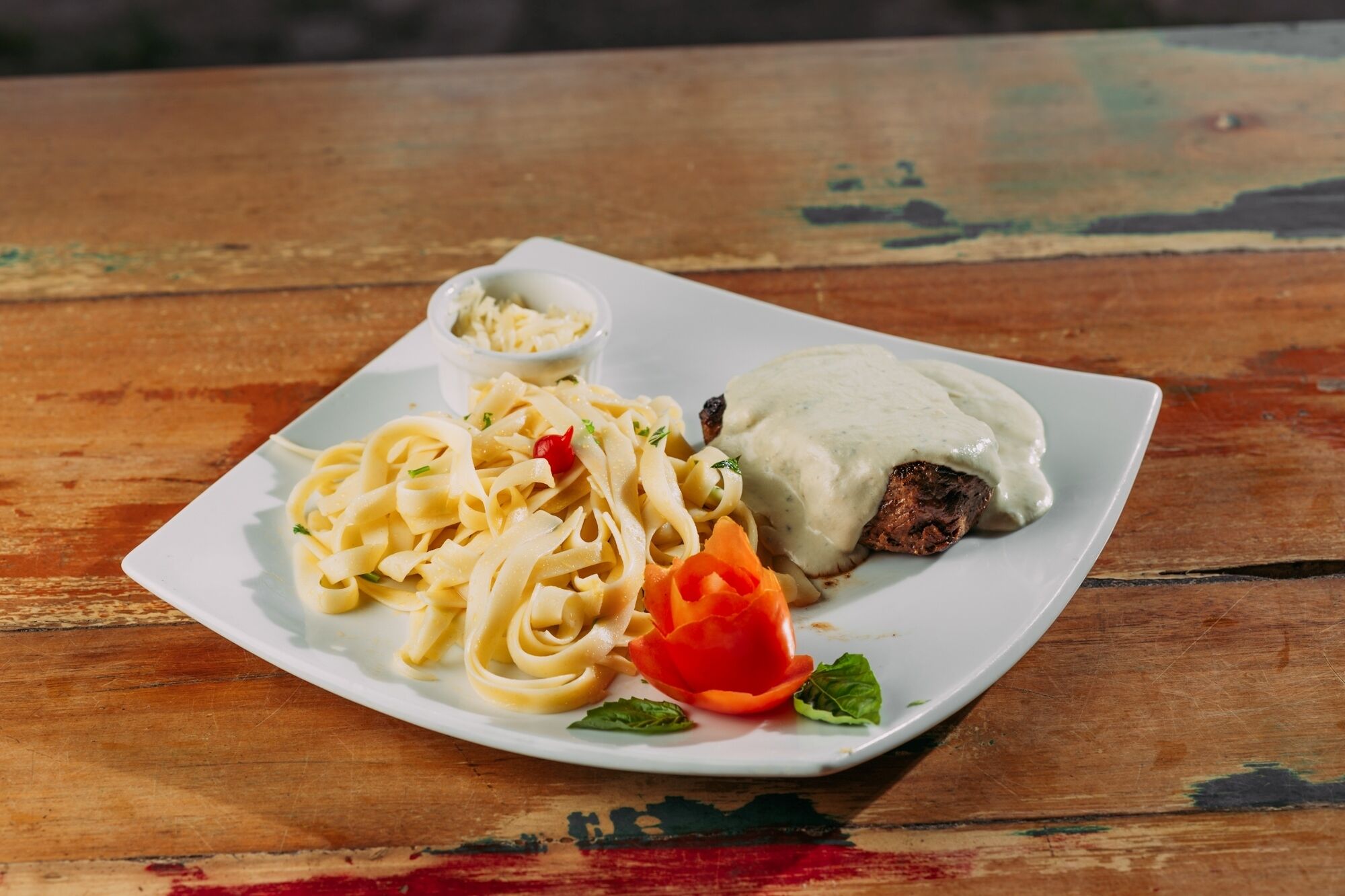 A plate of pasta with a meat patty and sauce, and a small bowl of sauce on a wooden table.