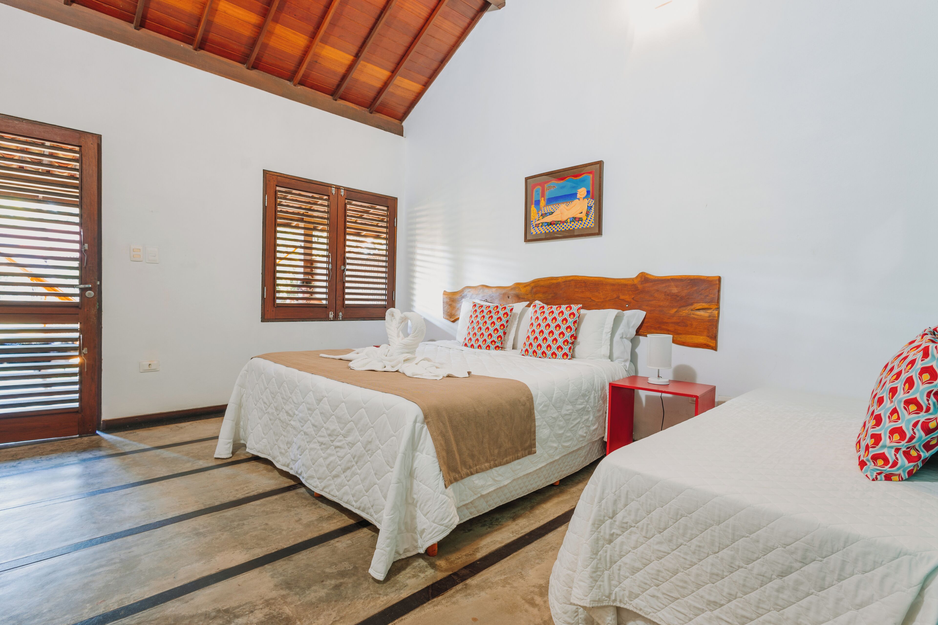 Une chambre d’hôtel avec deux lits, une tête de lit en bois, un tableau au mur et une table de chevet rouge.