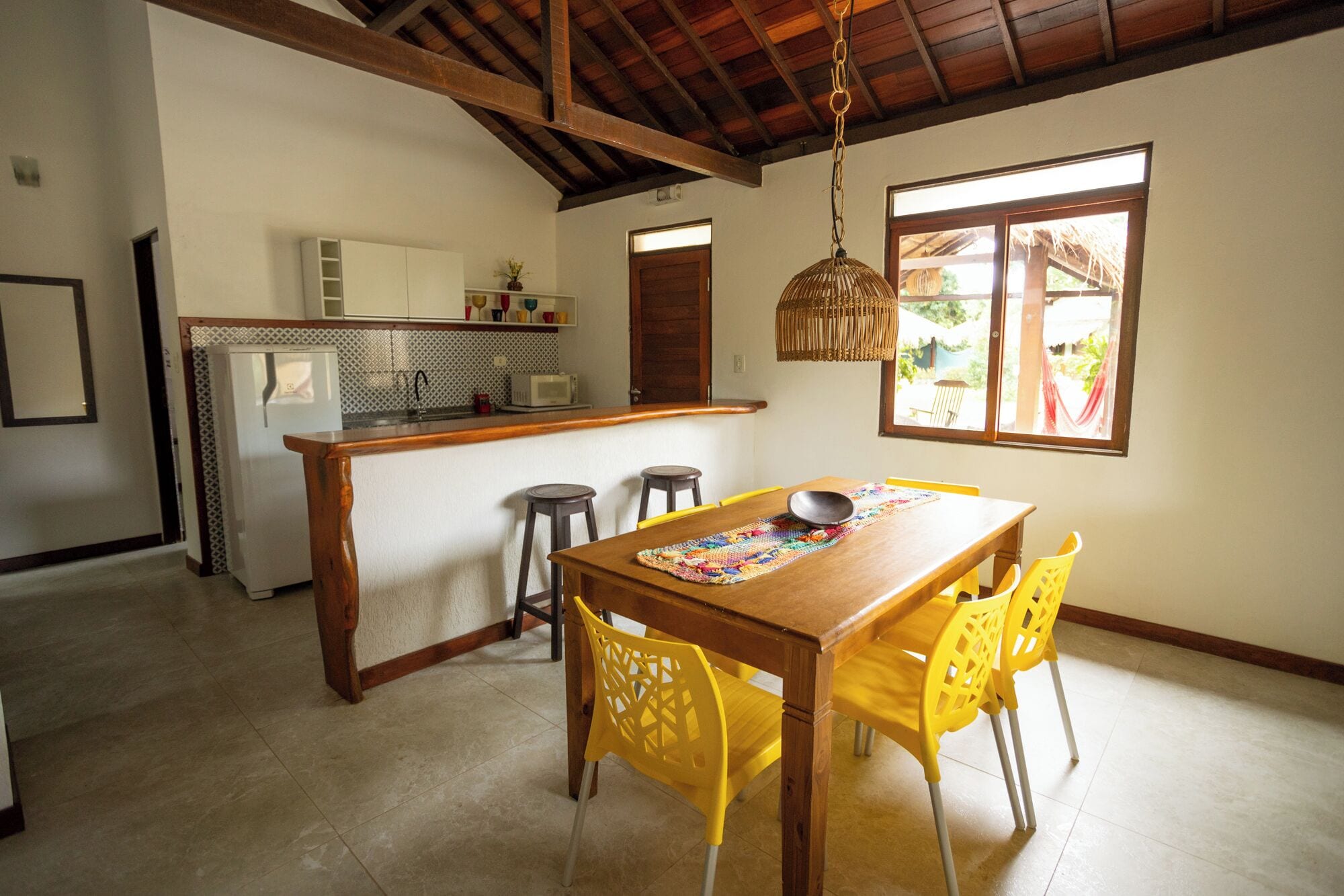 A dining area with a wooden table and yellow chairs, a kitchen with white cabinets, and a hanging light fixture.