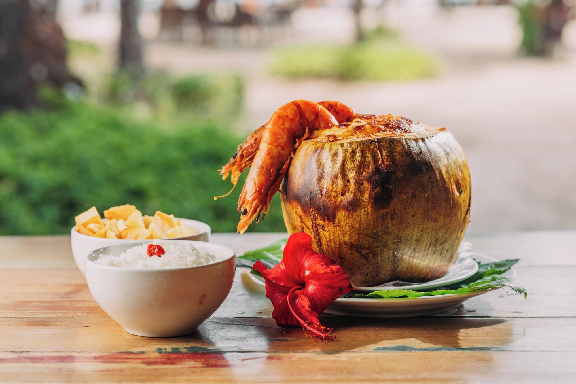 A coconut with a cooked shrimp on top, a bowl of rice, and a red flower on a wooden table.