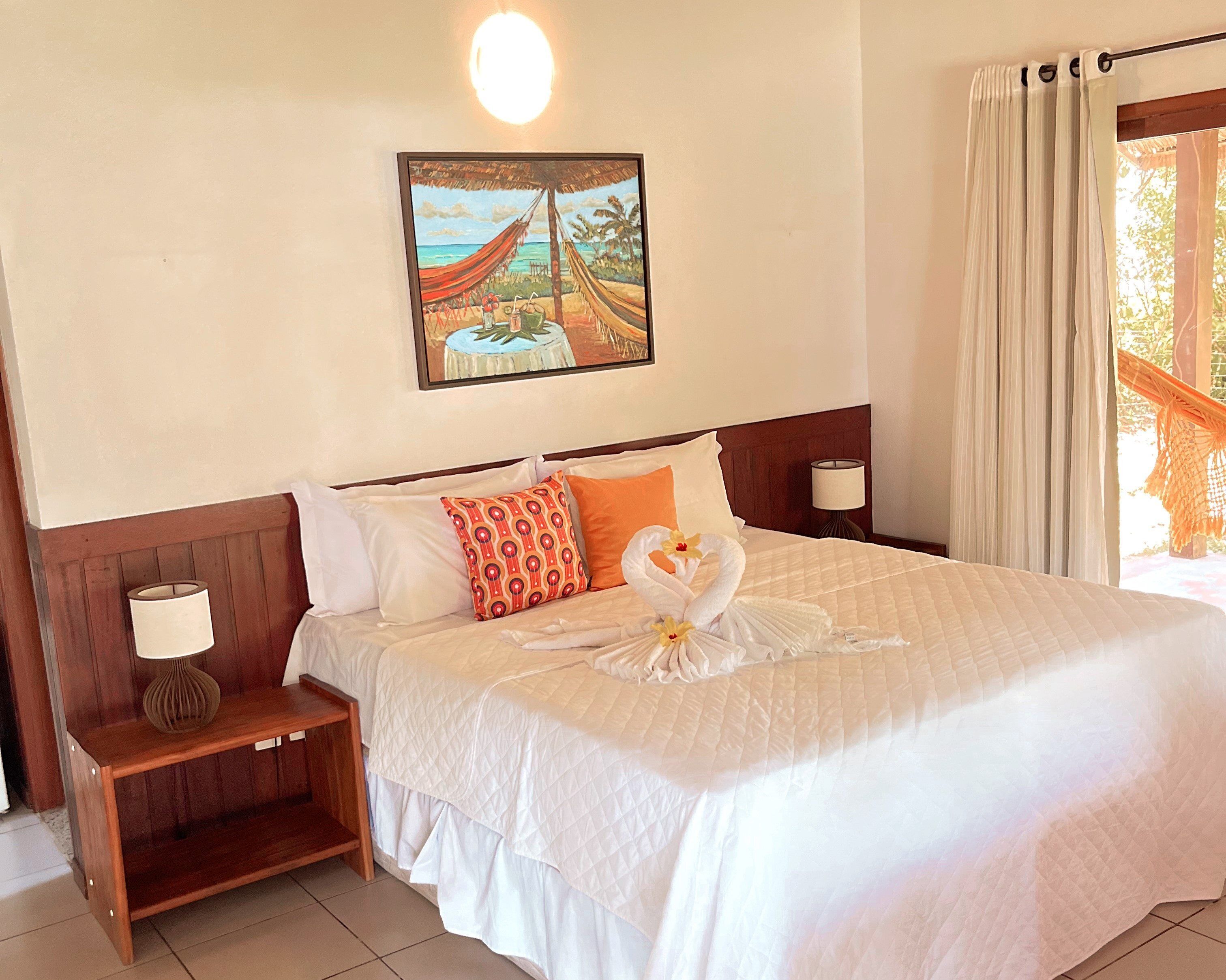 A neatly made bed with a quilted white bedspread, a wooden nightstand with a lamp, and a painting of a beach scene above the bed.