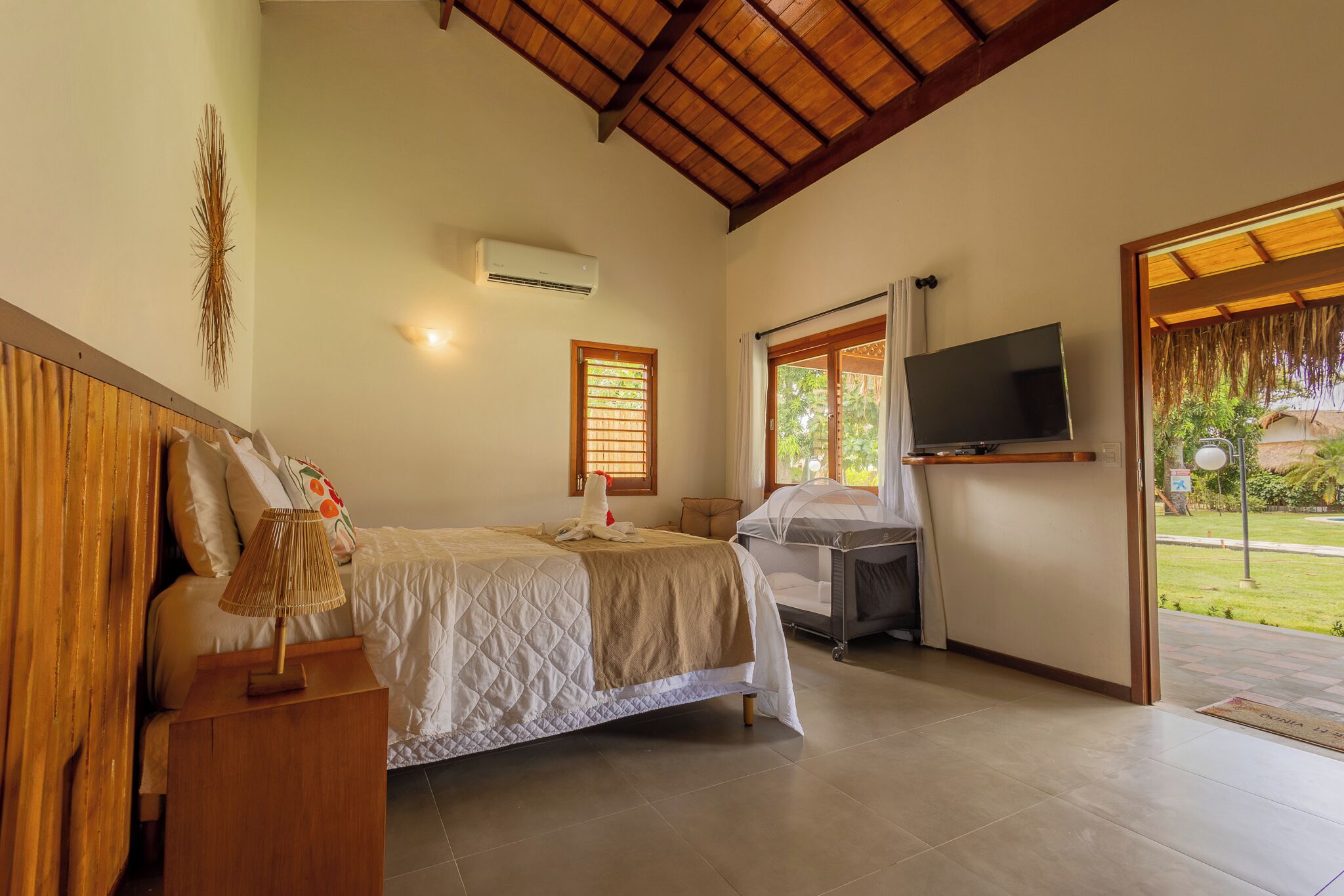 A bedroom with a bed, bedside table, TV, and a window with a view of greenery.
