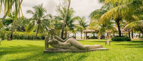 A sculpture of a reclining figure in a park with palm trees and a thatched-roof structure in the background.