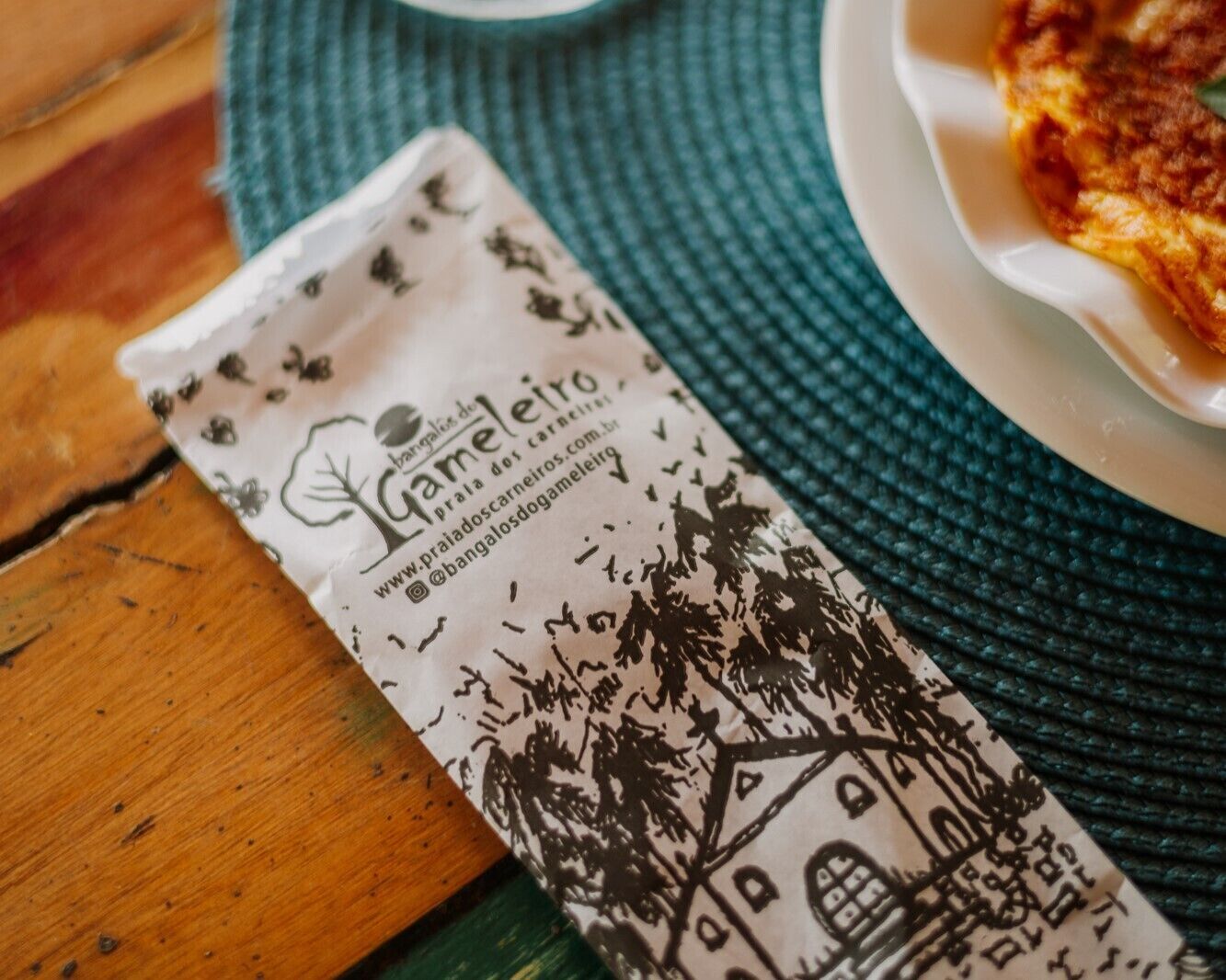 A packet of 'Café do Leir' coffee with a website and social media handle on it, placed on a wooden table next to a plate with food.