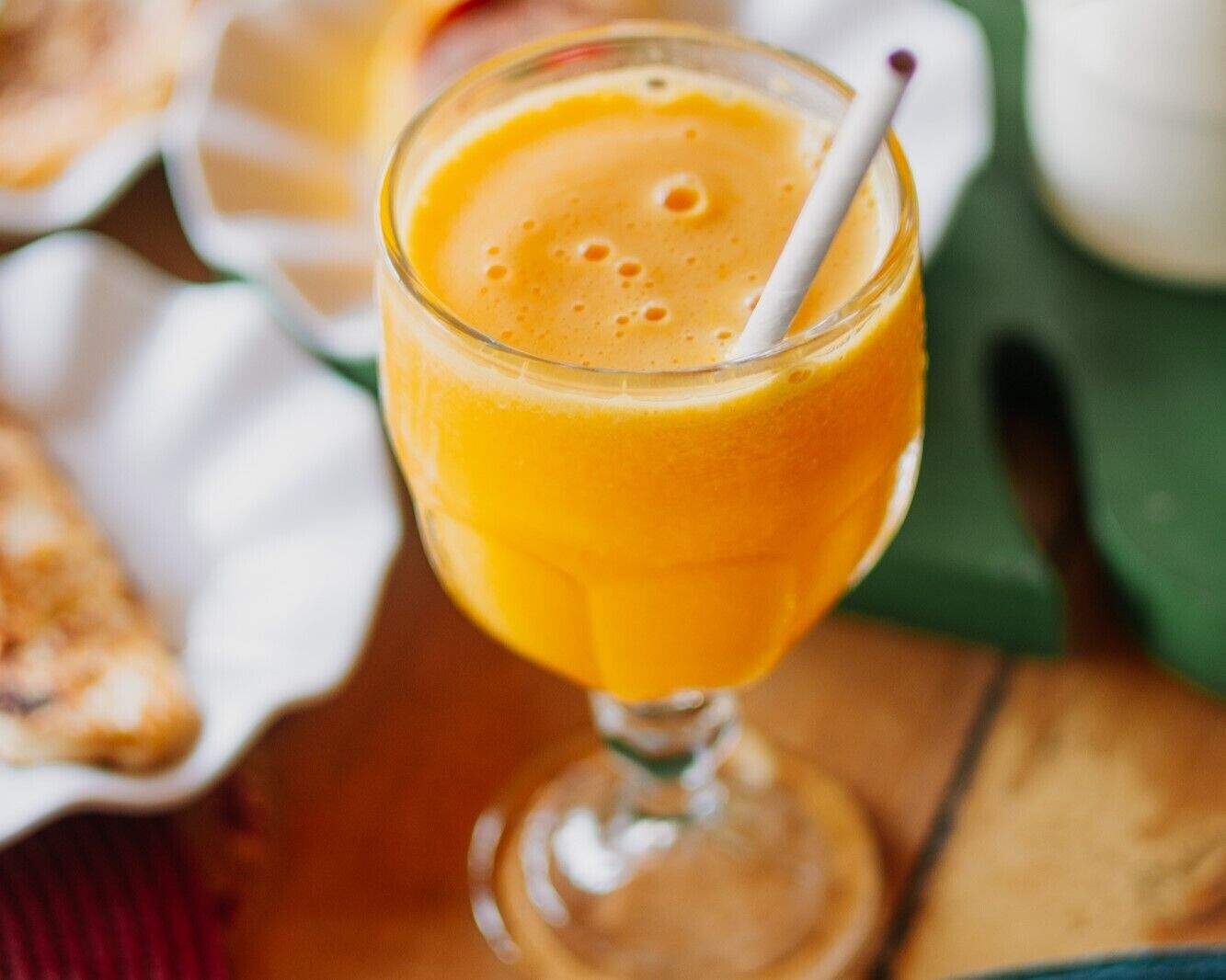 A glass of orange juice with a straw, placed on a wooden surface with a white plate and a green napkin in the background.