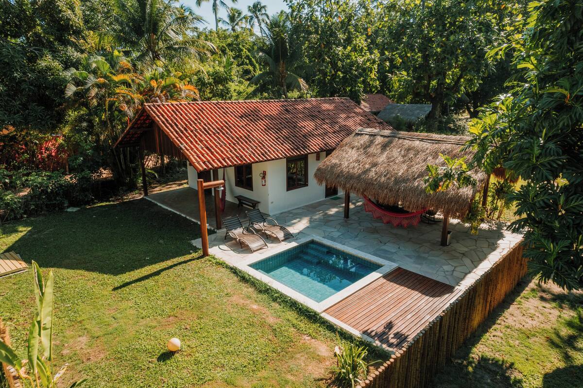 Une maison avec une piscine, entourée d’une végétation luxuriante et dotée d’une structure à toit de chaume.