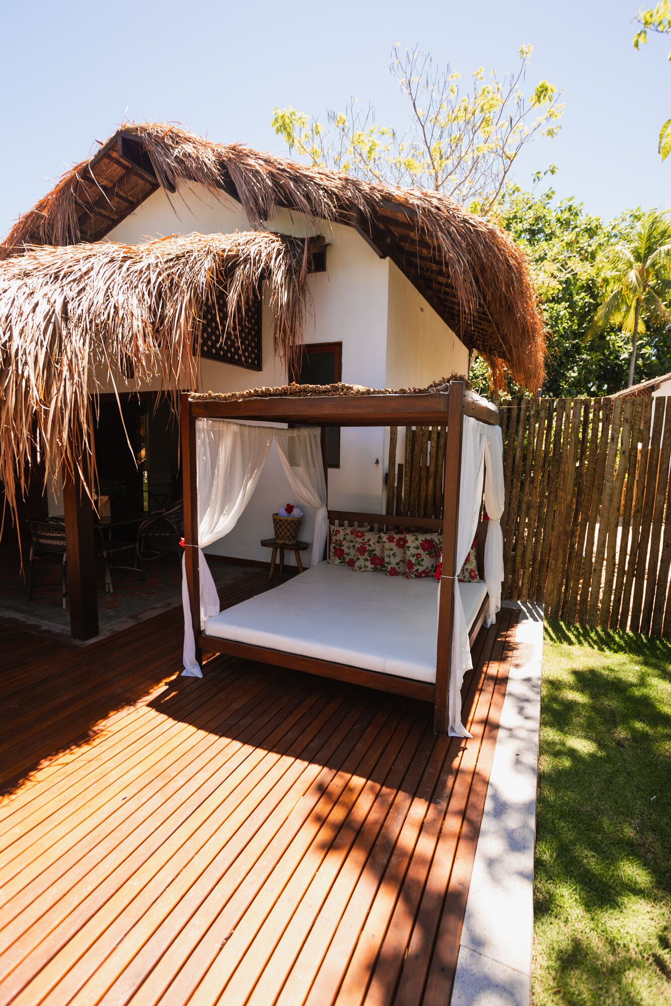 A thatched-roof outdoor sleeping area with a wooden bed frame, a small table, and a chair.