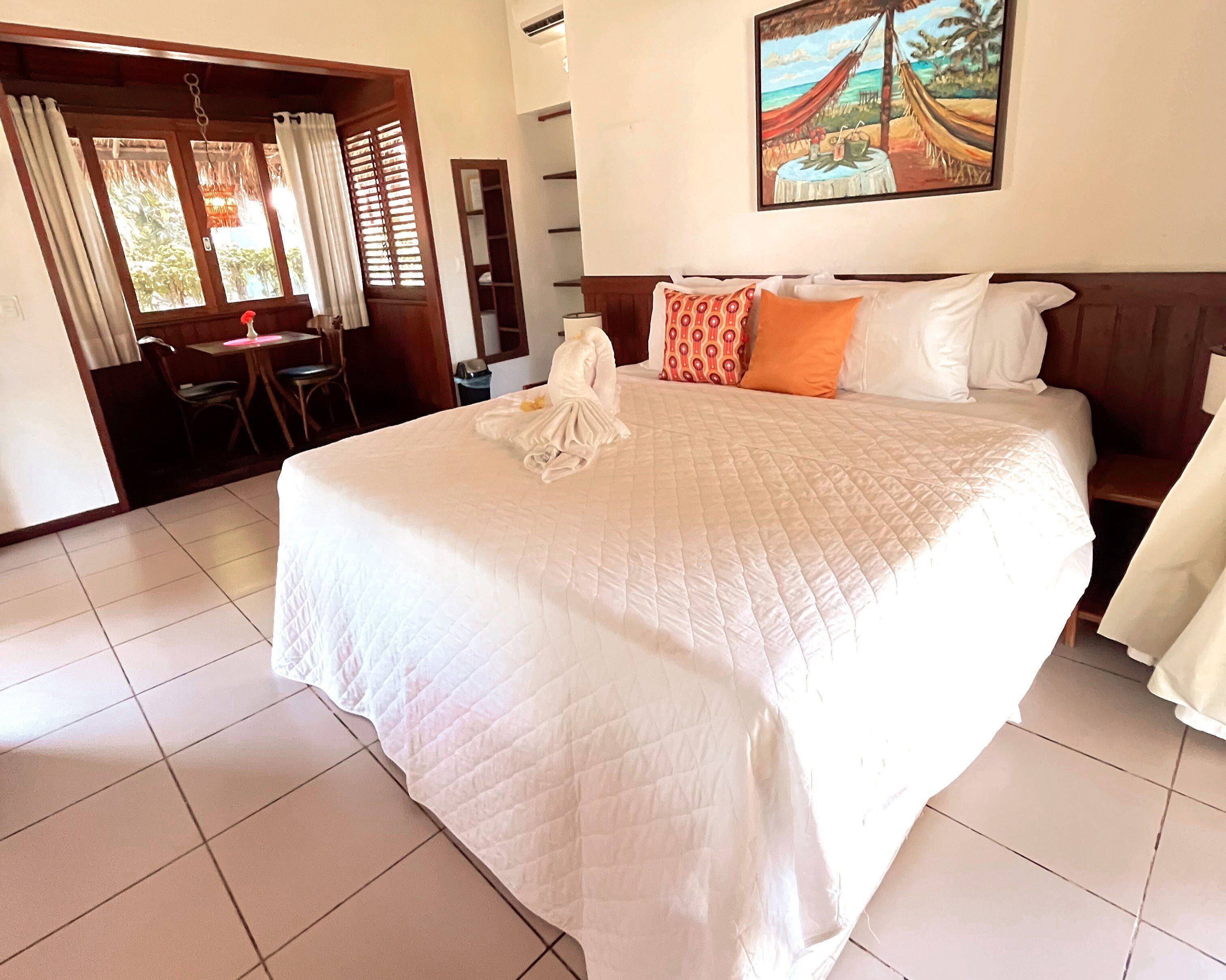Habitación de hotel con una cama grande, un cuadro de una playa, una zona de comedor con mesa y sillas, y vista al exterior a través de una ventana con cortinas.