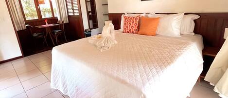 Habitación de hotel con una cama grande, un cuadro de una playa, una zona de comedor con mesa y sillas, y vista al exterior a través de una ventana con cortinas.