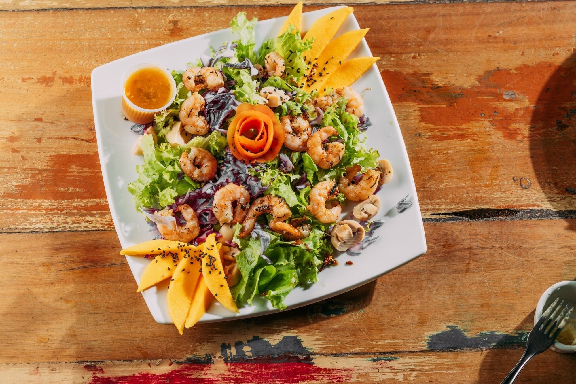 A square plate with a salad, shrimp, and mango slices on a wooden table.