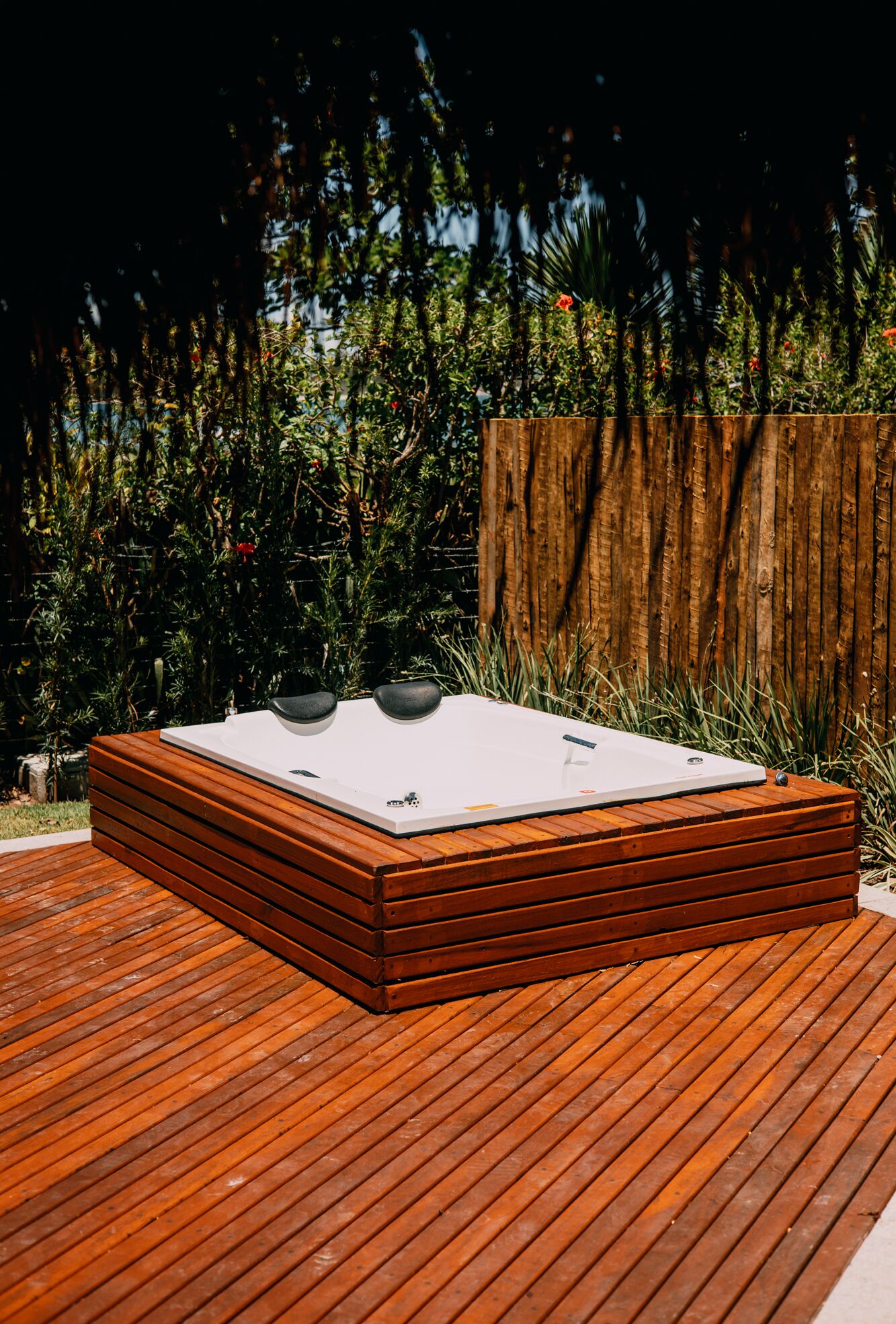 Una vasca idromassaggio su un pontile di legno, con un recinto in legno e vegetazione sullo sfondo.