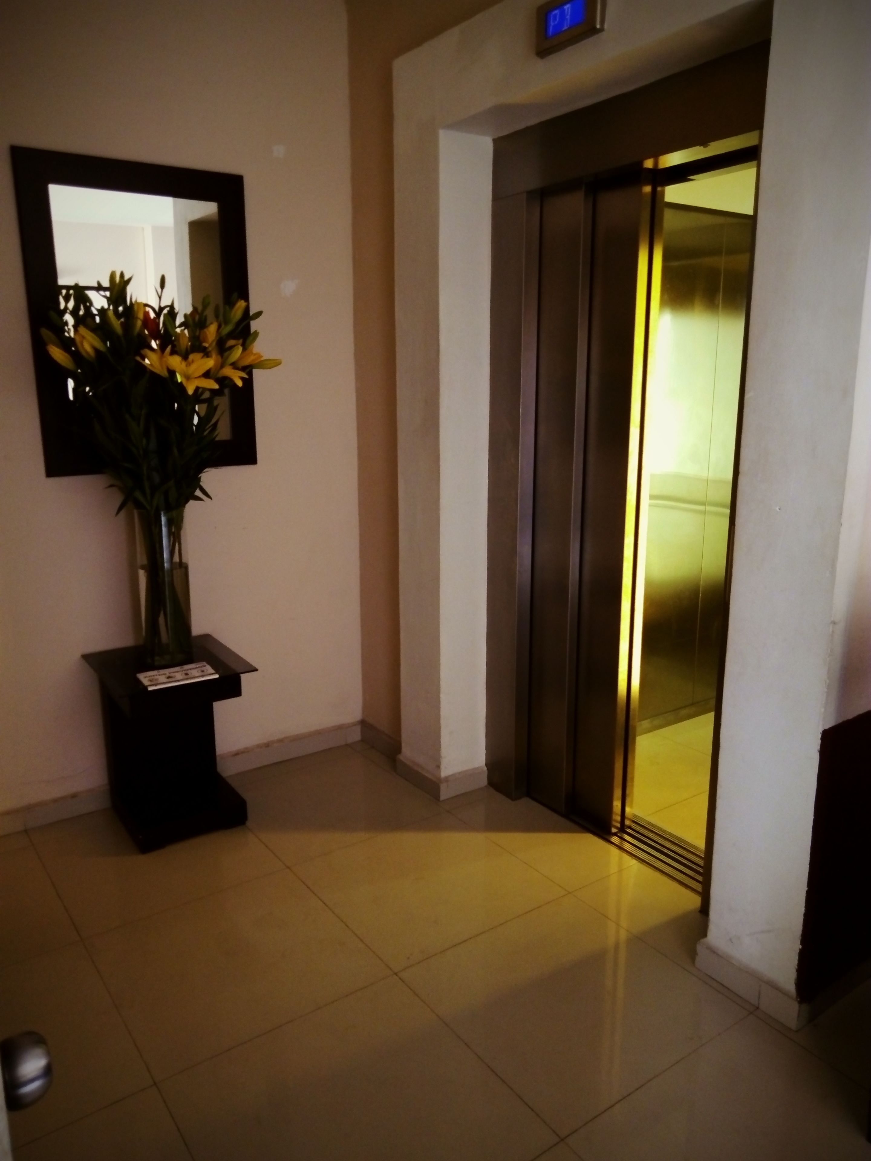 Un hall con un jarrón de flores amarillas sobre un pedestal, un espejo en la pared y un ascensor con pantalla digital.