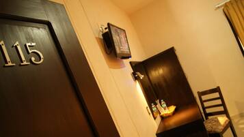 A hotel room with a door labeled '115', a mounted television, a dark wooden cabinet, and a chair with a patterned cushion.