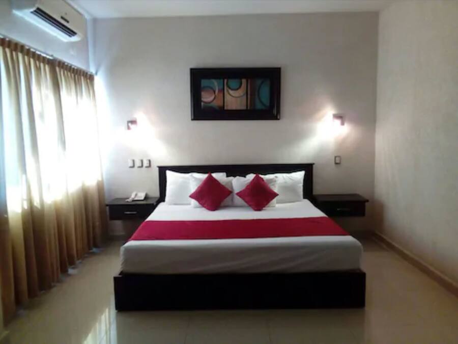 A hotel room with a large bed, two bedside tables, a nightstand, a painting on the wall, and a window with curtains.