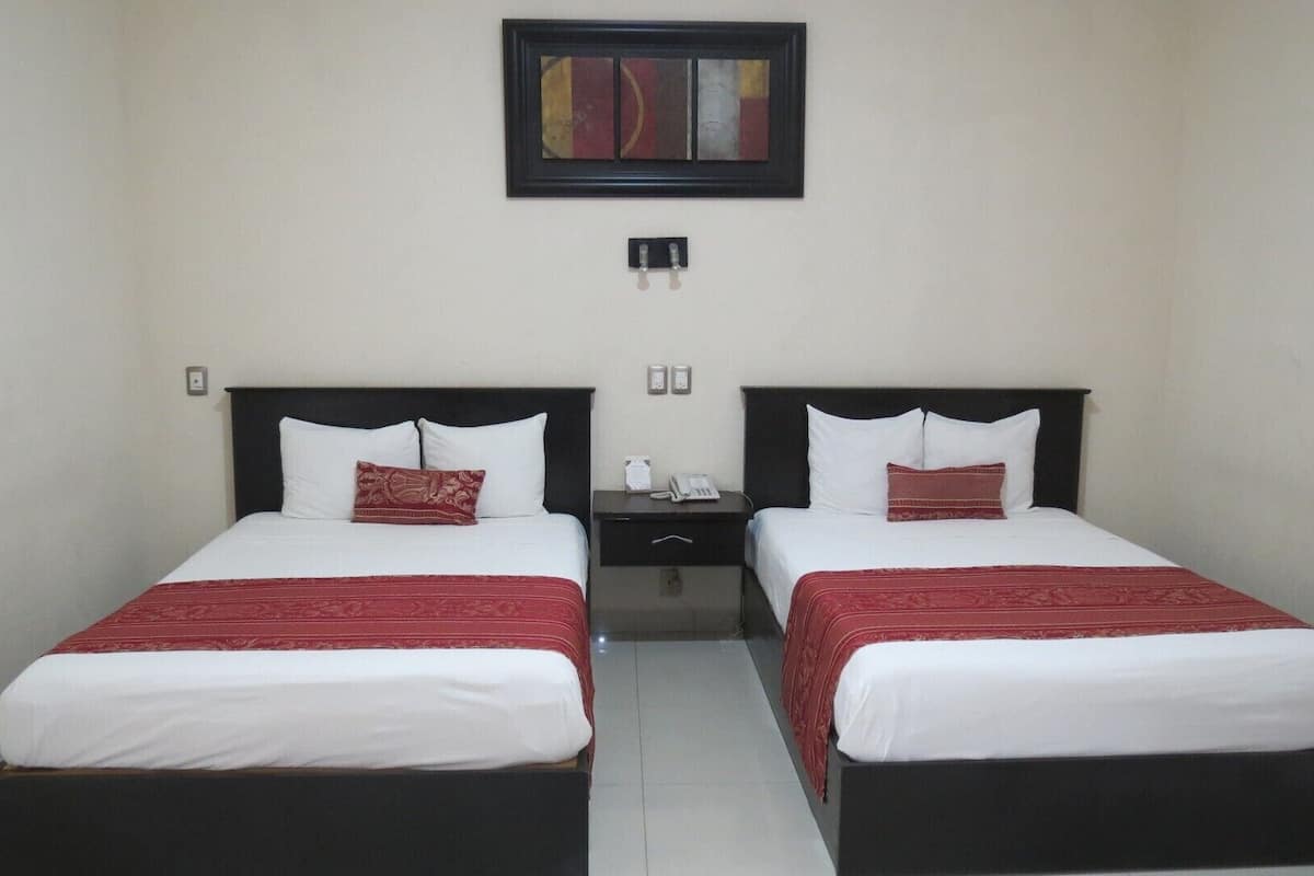 A hotel room with two beds, each with a black headboard, white bedding, and a red accent pillow. There is a nightstand between the beds with a phone and a small table. Above the beds, there is a framed artwork on the wall.