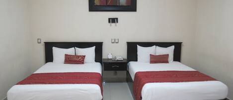 A hotel room with two beds, each with a black headboard, white bedding, and a red accent pillow. There is a nightstand between the beds with a phone and a small table. Above the beds, there is a framed artwork on the wall.