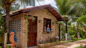 A small brick house with a wooden door and window, surrounded by lush greenery and decorative sculptures.