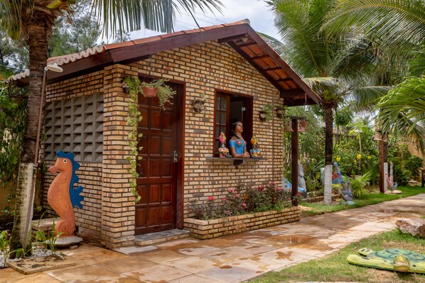 Uma pequena casa de tijolos com porta e janela de madeira, rodeada por vegetação exuberante e esculturas decorativas.