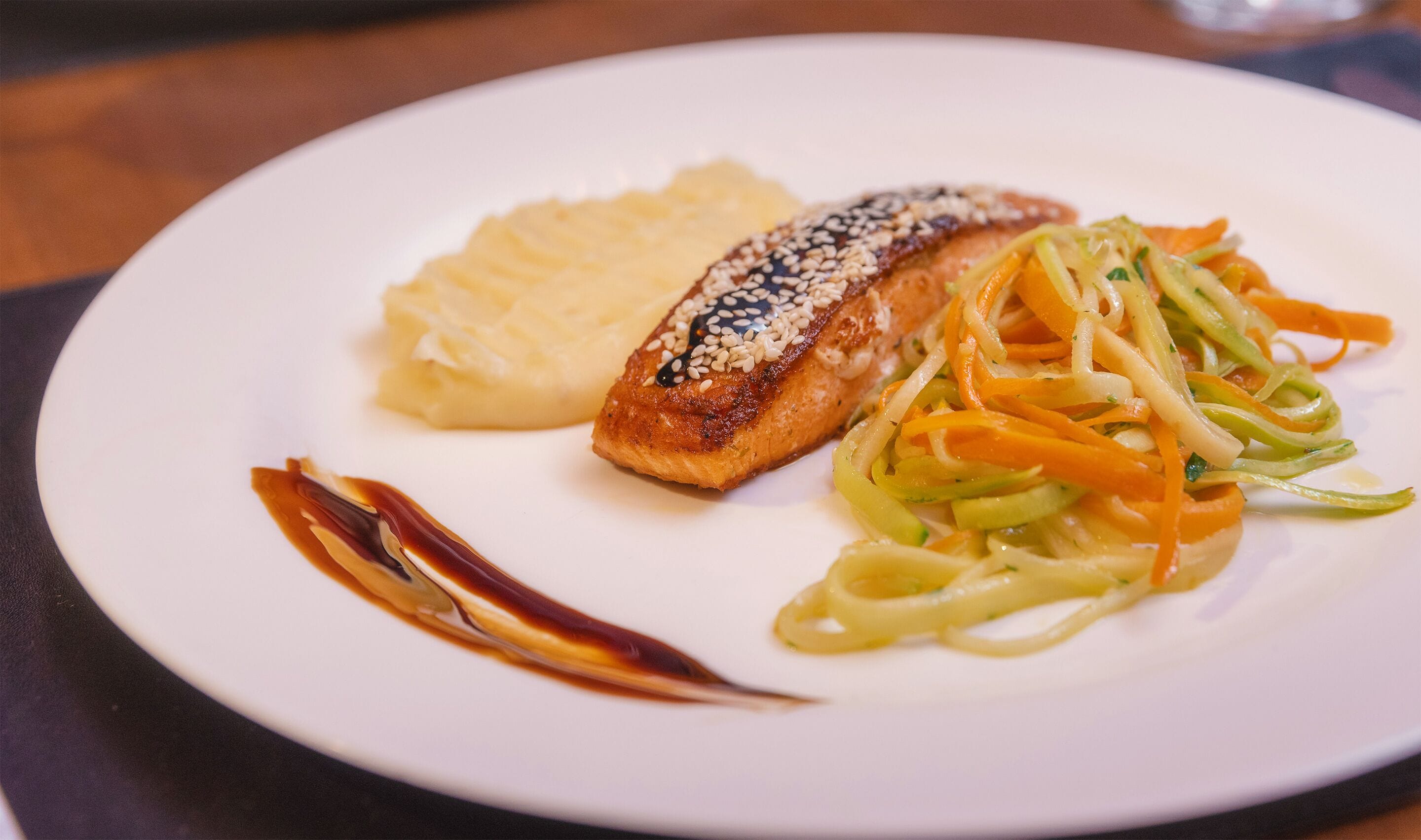 Un plato con un trozo de salmón cubierto de semillas de sésamo, puré de papas y una guarnición de verduras salteadas.