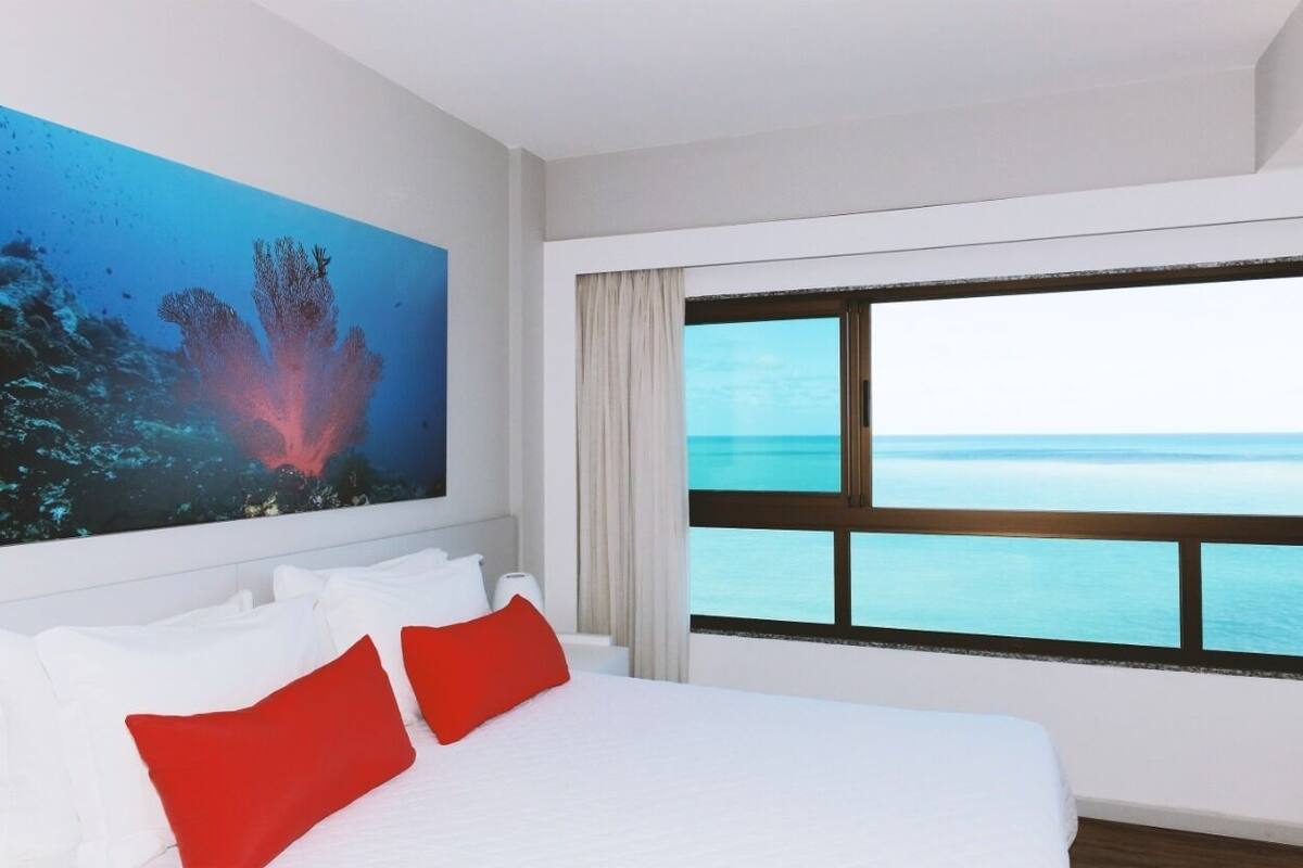 A hotel room with a large bed, a painting of an underwater scene, and a view of the ocean through a large window.
