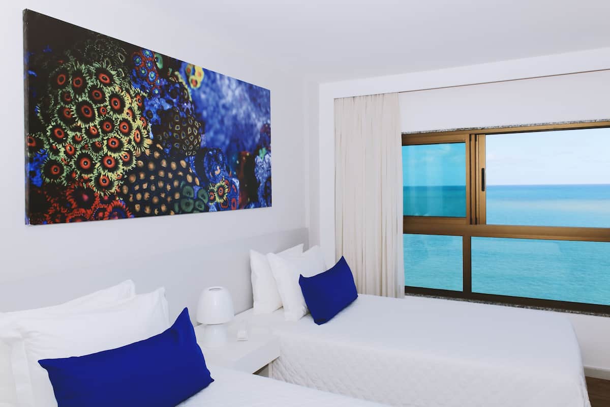 A hotel room with a large window offering a view of the sea, two beds with white bedding and blue pillows, a white nightstand, and a colorful abstract painting on the wall.