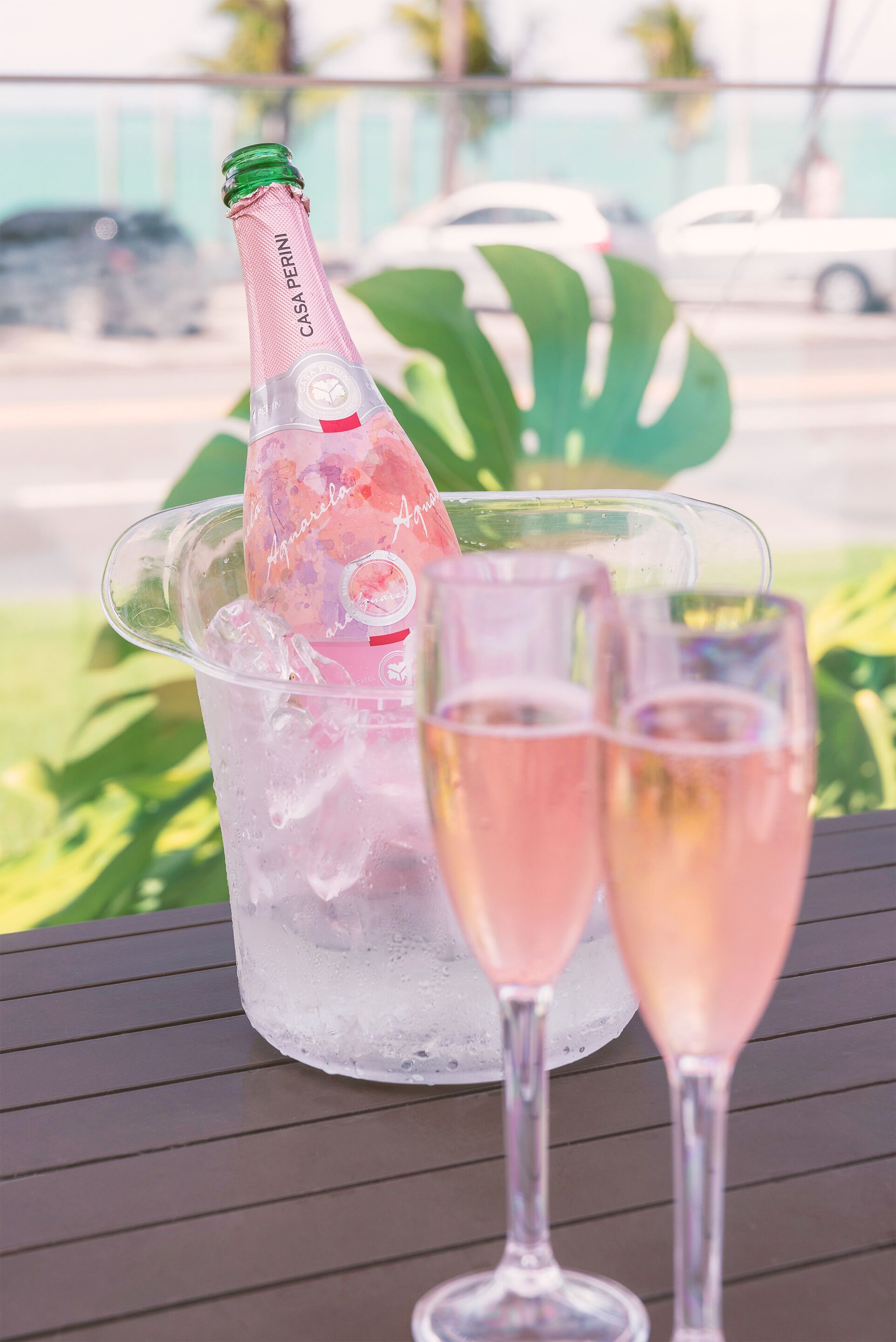 Una botella de champán Casa Perini en un cubo de hielo transparente con dos copas llenas de champán rosado.