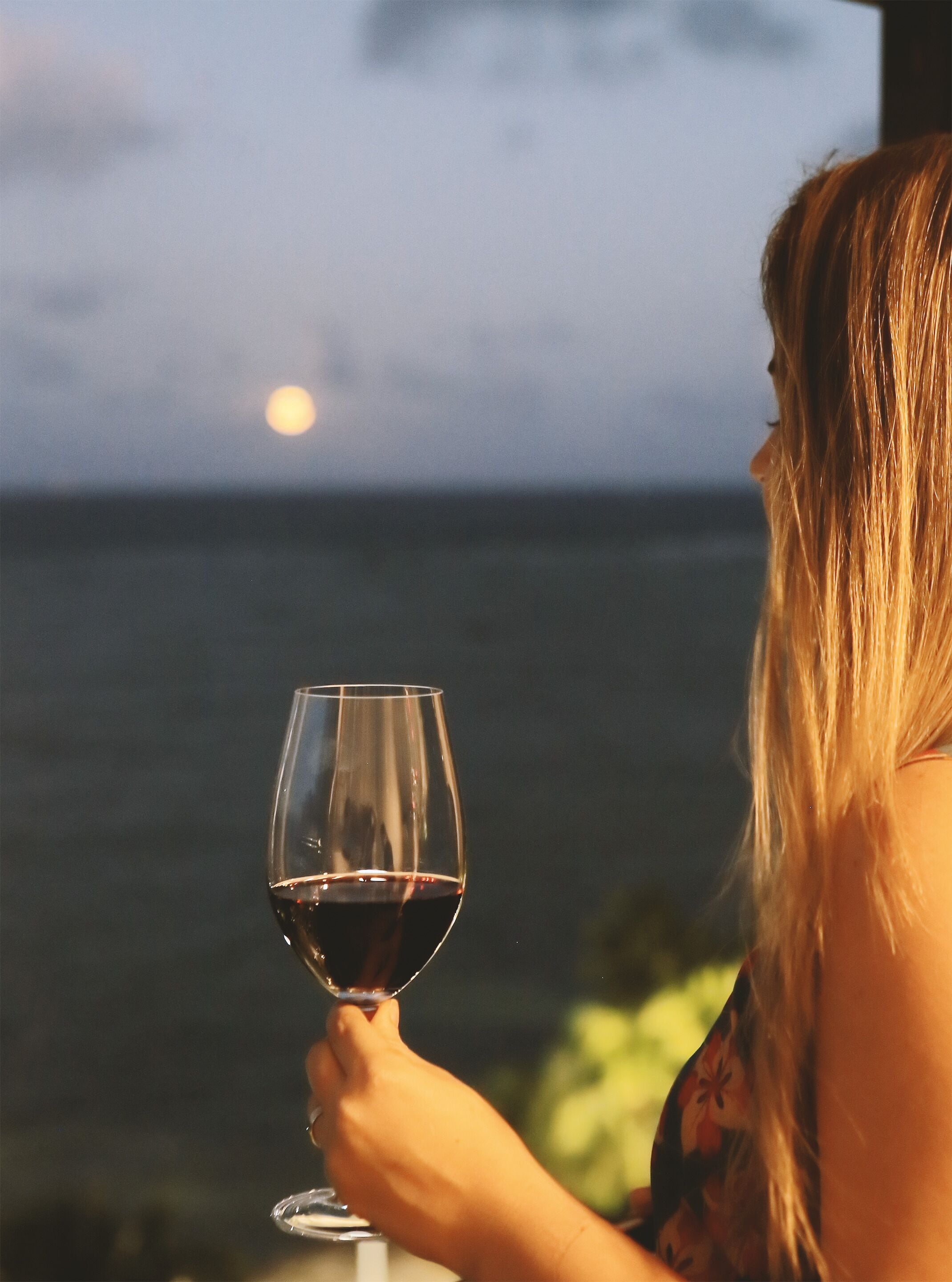 Una persona sosteniendo una copa de vino con vista al océano y una luna de fondo.