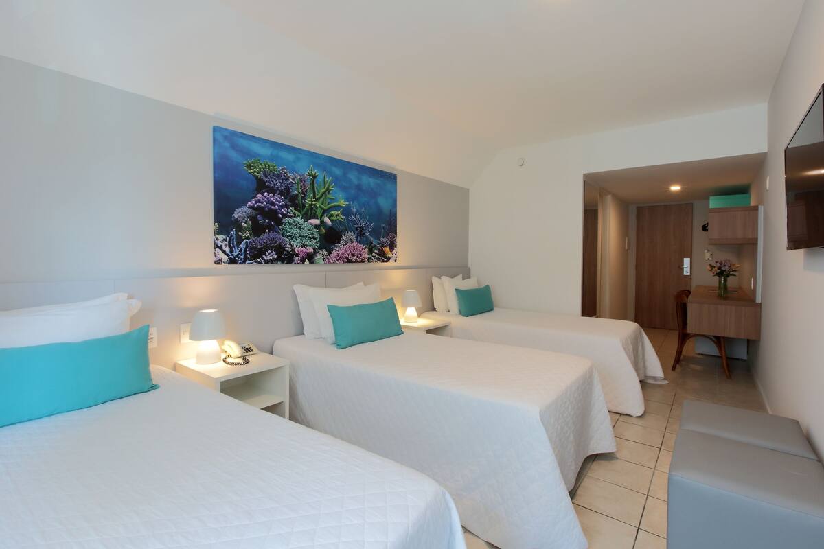 A hotel room with two beds, a painting of an underwater scene, a desk with a chair, and a TV mounted on the wall.