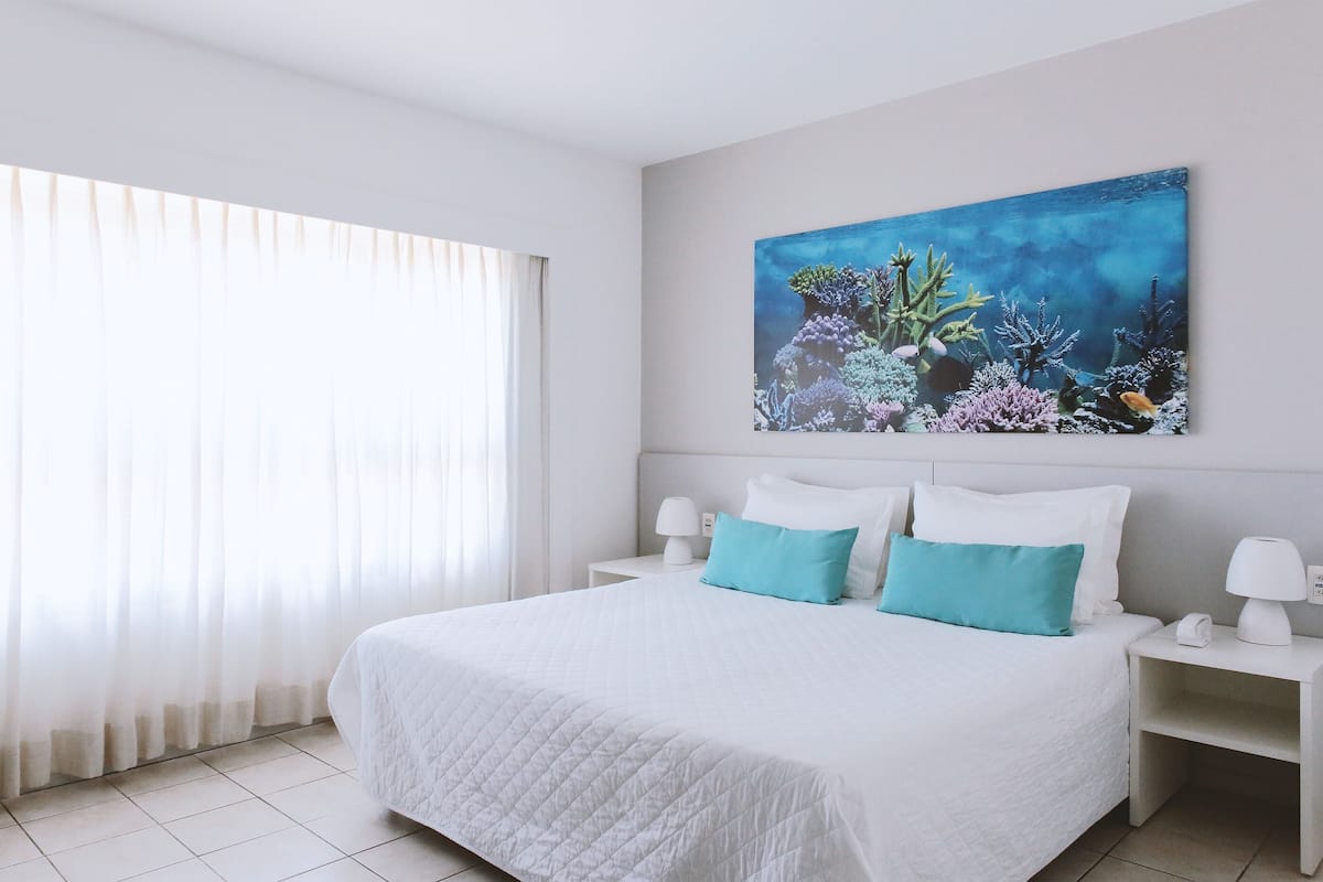 A neatly made bed with white bedding and two teal pillows, a white nightstand with a lamp, and a wall-mounted painting of an underwater scene.