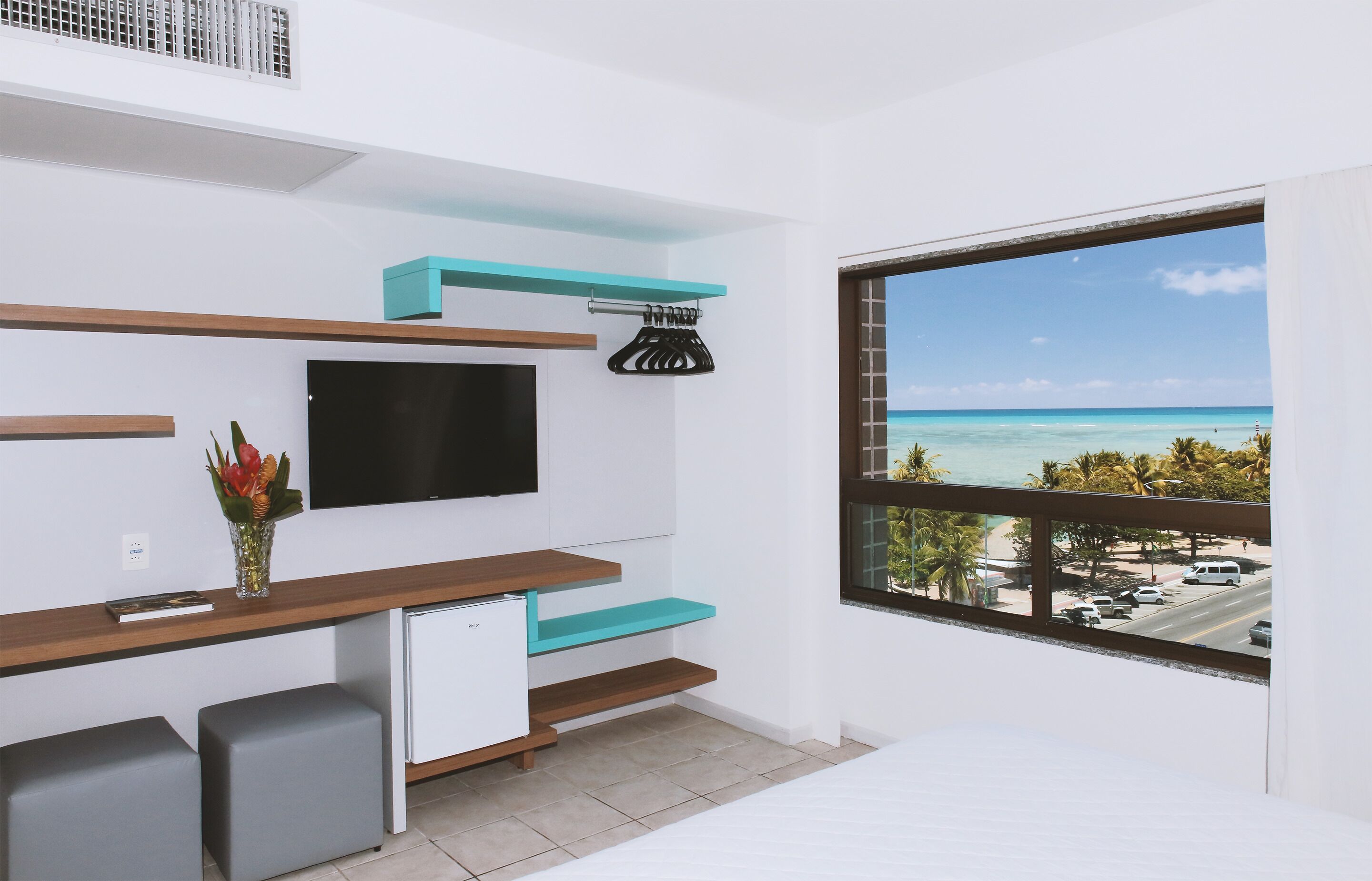 A modern room with a TV, a small fridge, a desk with a chair, a window with a view of the beach and ocean, and a bed.