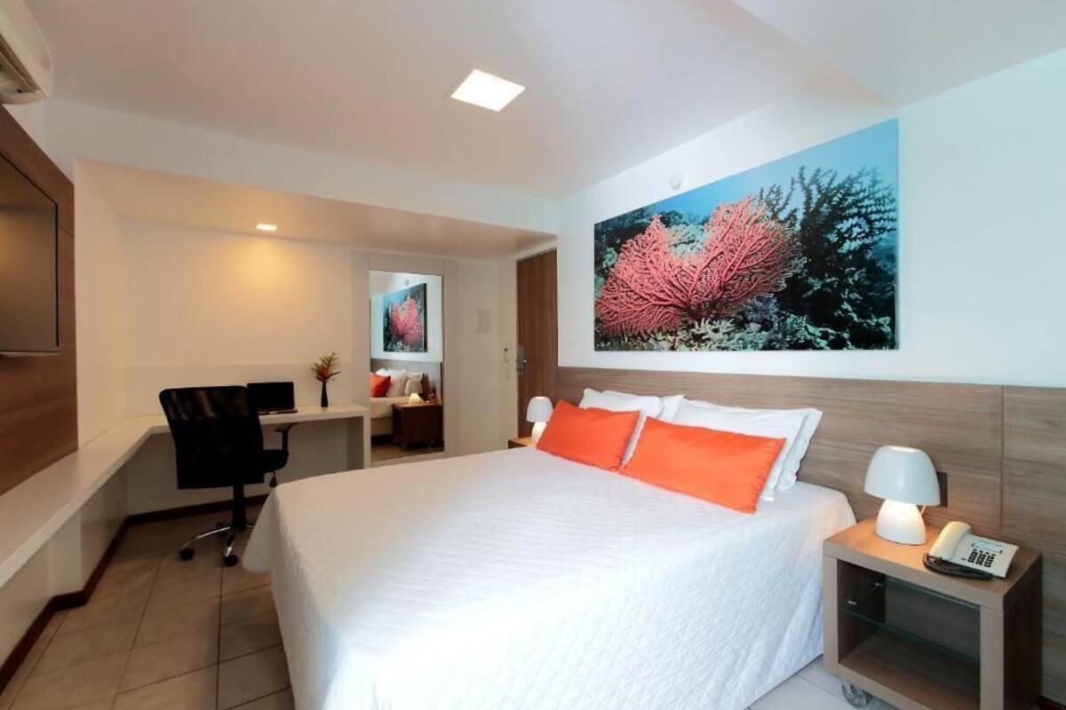 A hotel room with a large bed, a desk, a chair, a nightstand with a lamp, and a painting on the wall.