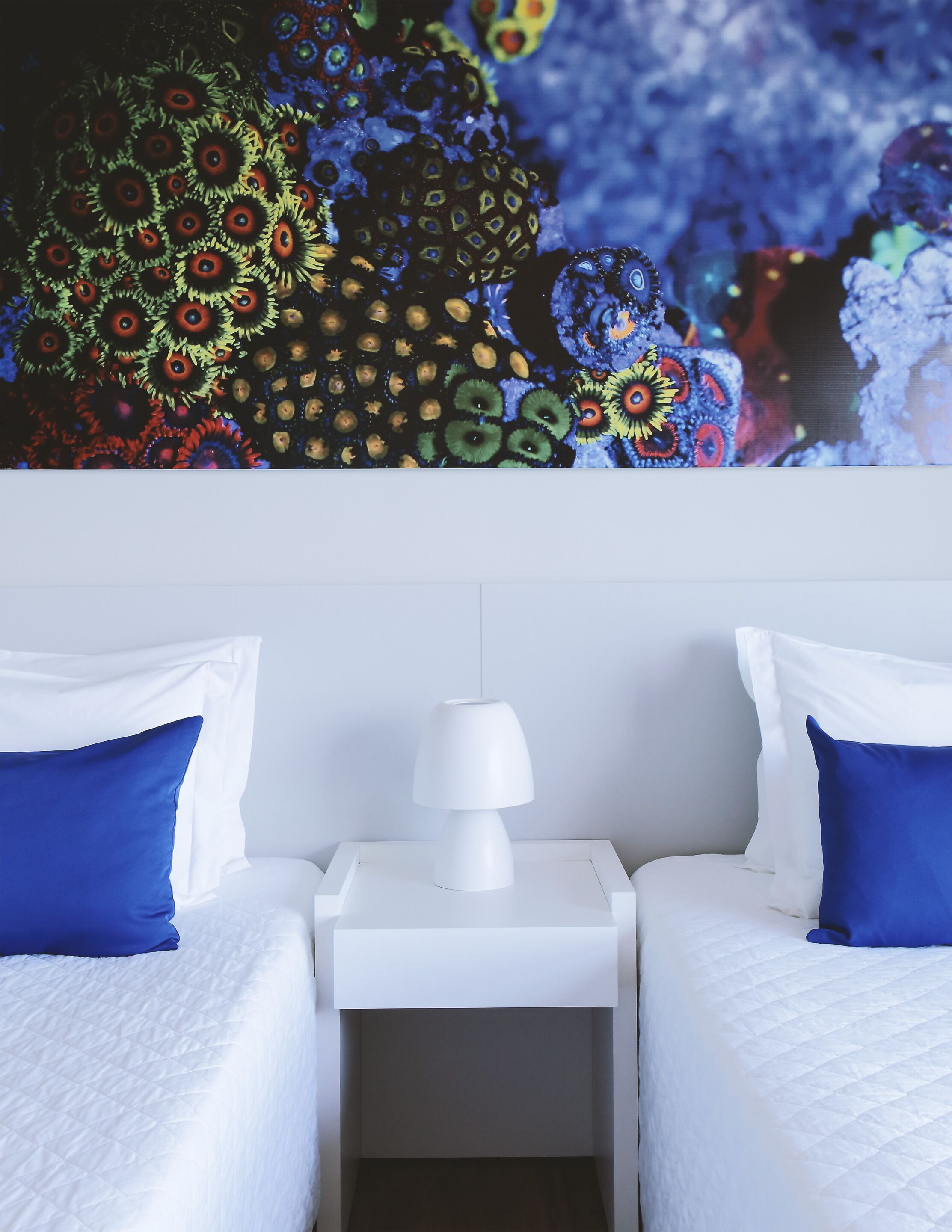 A hotel room with two beds, white bedding, a white nightstand with a lamp, and a colorful mural on the wall.