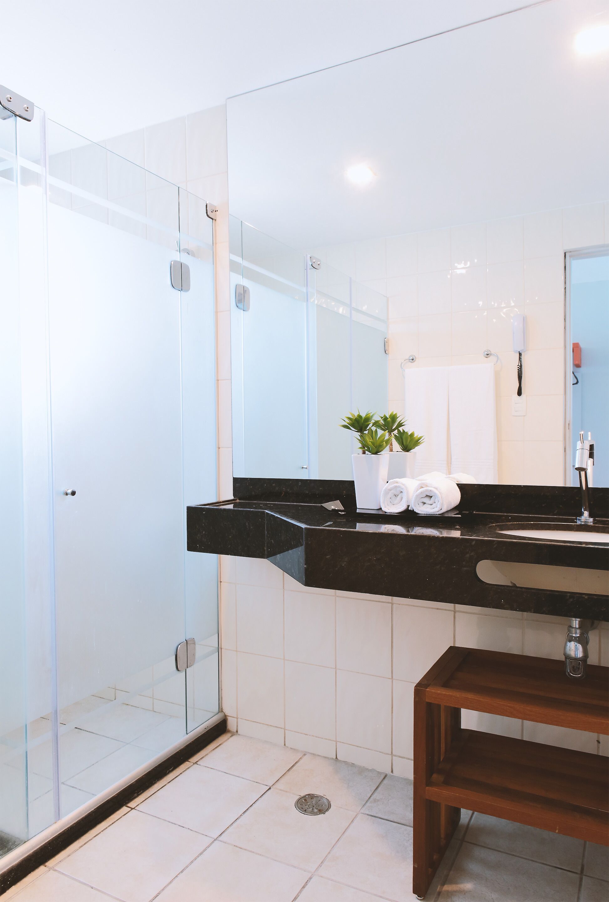 Un baño moderno con cabina de ducha de vidrio, encimera negra, banco de madera y suelo de baldosas blancas.